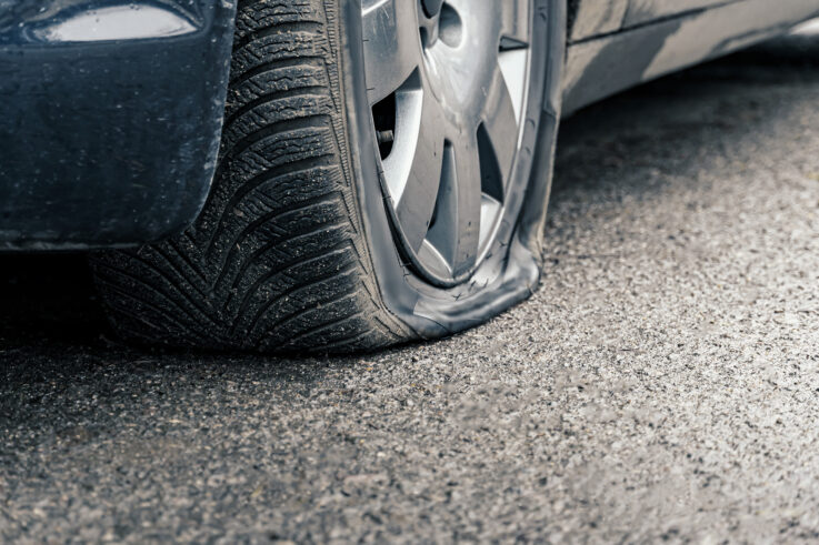 A car is shown with a completely flat tire, the rubber compressed against the pavement. The tire appears to have lost all air pressure, suggesting the need for repair or replacement.