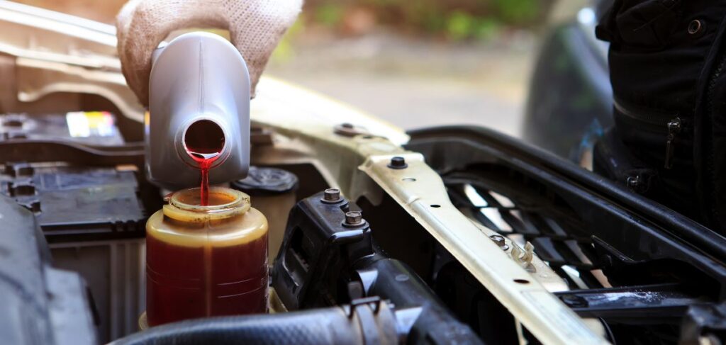 Person pouring red power steering fluid from a bottle into a reservoir under the hood of a car.