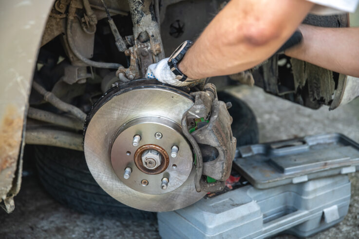 A close-up of a person repairing a car brake system, showing their hands working on the exposed brake rotor and caliper. The vehicle is raised, and a toolbox is placed nearby on the ground.