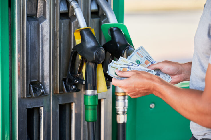 Cropped view of man holding hundred-dollar bills at a gas station pump.