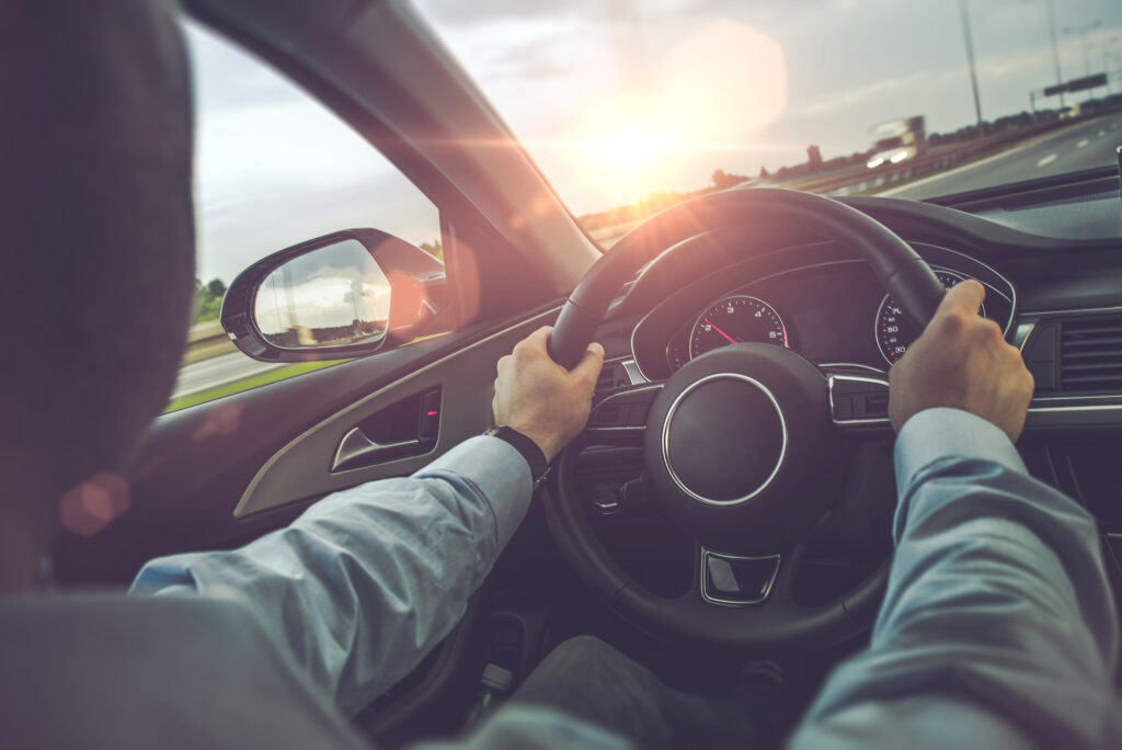 A person is driving a car on a highway at sunset with both hands on the steering wheel and the dashboard in view. The sun is low on the horizon, casting a warm glow through the windshield.