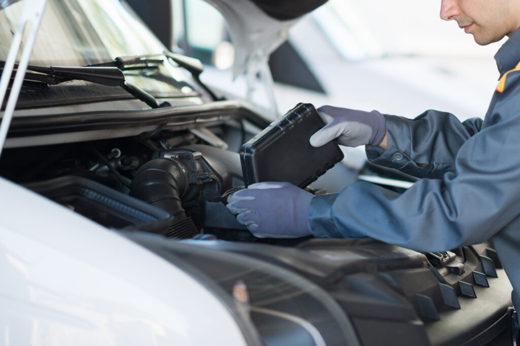  A mechanic wearing gloves works on a vehicle engine, inspecting or replacing a black rectangular component under the hood.