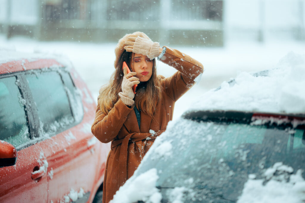 A frustrated woman in winter clothing stands outside in a snowy parking lot, talking on her phone while brushing snow off her forehead and a snow-covered car.