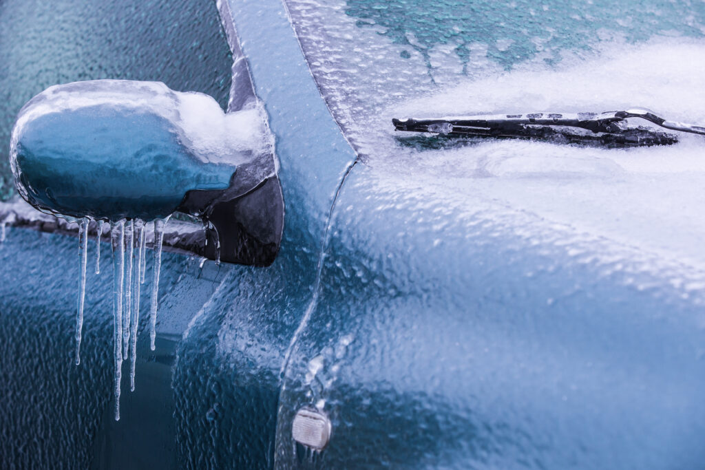 A close-up of a car covered in a thick layer of ice with icicles hanging from the side mirror and frozen windshield wipers.