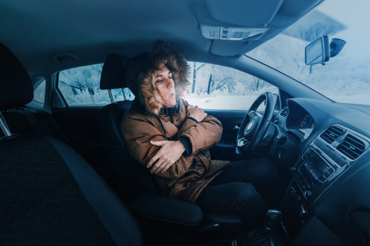 A person sitting inside a car on a snowy day, bundled in a thick coat with a fur-lined hood, crossing their arms and exhaling visible breath, suggesting the car heater isn't working.