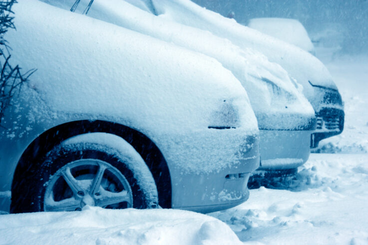 Several cars are parked outdoors, heavily covered in snow during a blizzard, illustrating severe winter weather conditions.